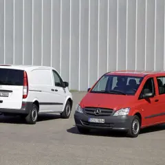 Dwa busy Mercedes-Benz: biały towarowy i czerwony osobowy, stojące na tle szarej ściany.