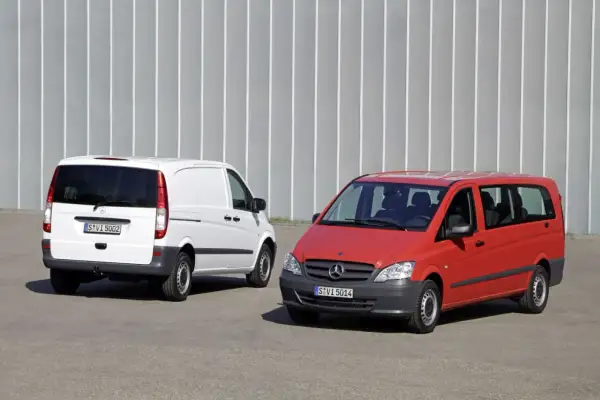Dwa busy Mercedes-Benz: biały towarowy i czerwony osobowy, stojące na tle szarej ściany.